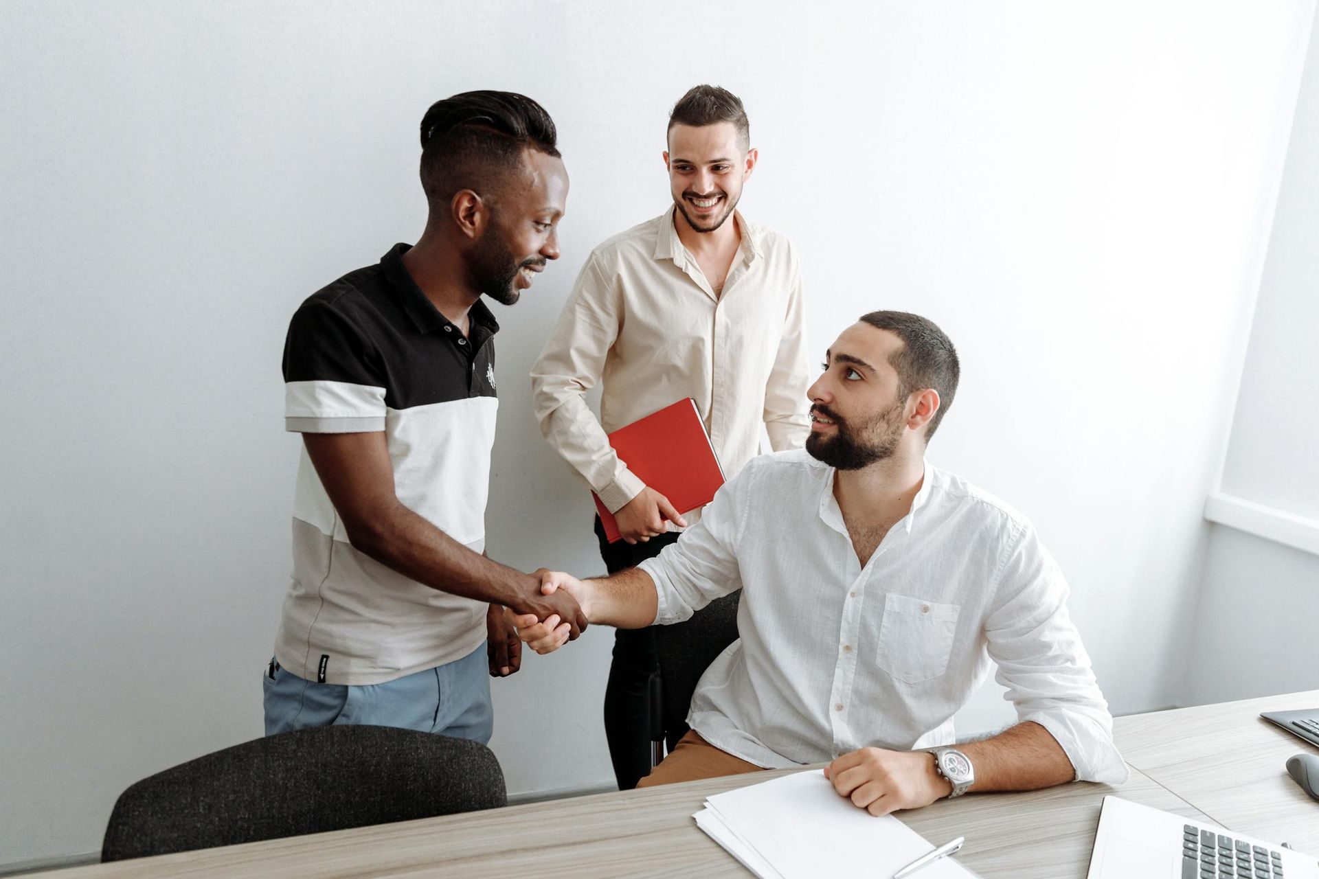 Three coworkers shake hands in an office, with one holding a red folder.