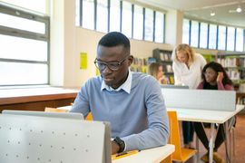 Student working on a laptop in a bright classroom, with two people studying at desks behind him.