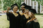 Four graduates in black caps and gowns taking a selfie outdoors, smiling in front of campus columns