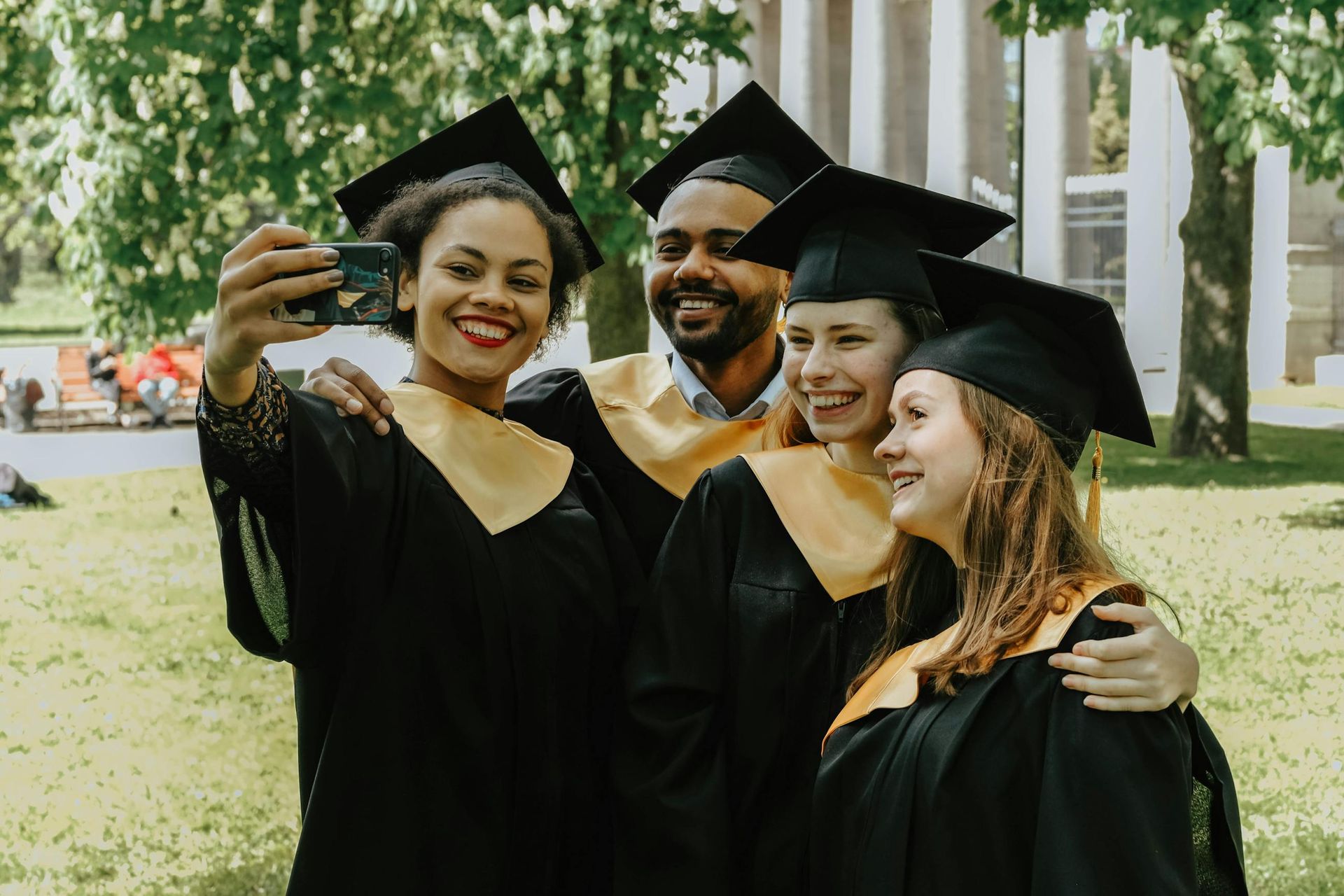 Four graduates in black caps and gowns taking a selfie outdoors, smiling in front of campus columns