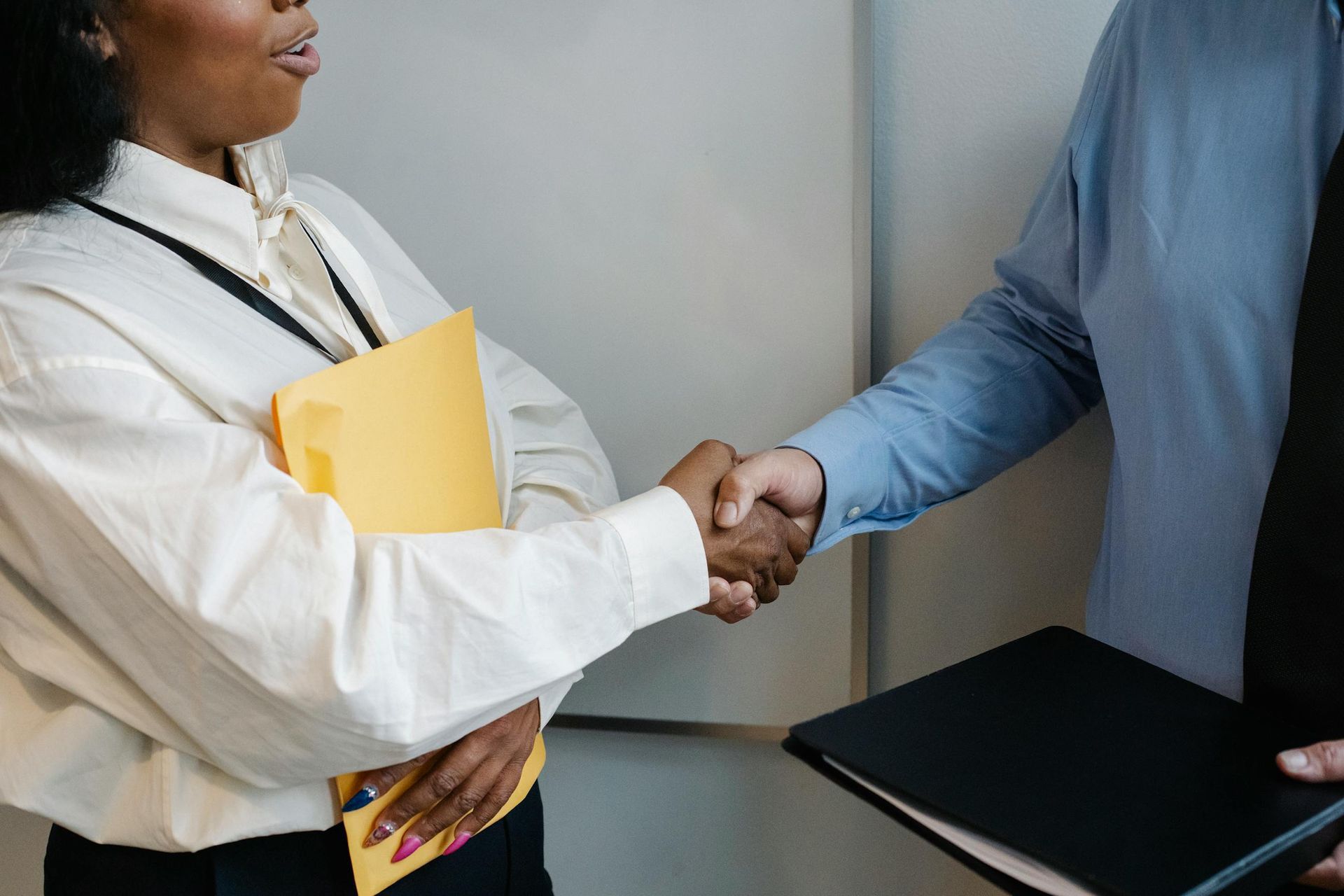 Two people shaking hands beside a desk, one holding a yellow folder