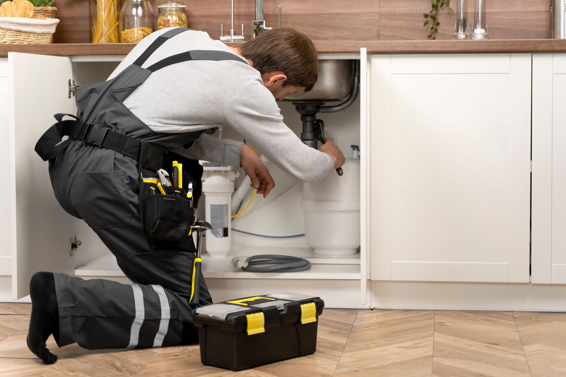 Plumber kneeling under a sink, repairing pipes with tools in a kitchen cabinet.