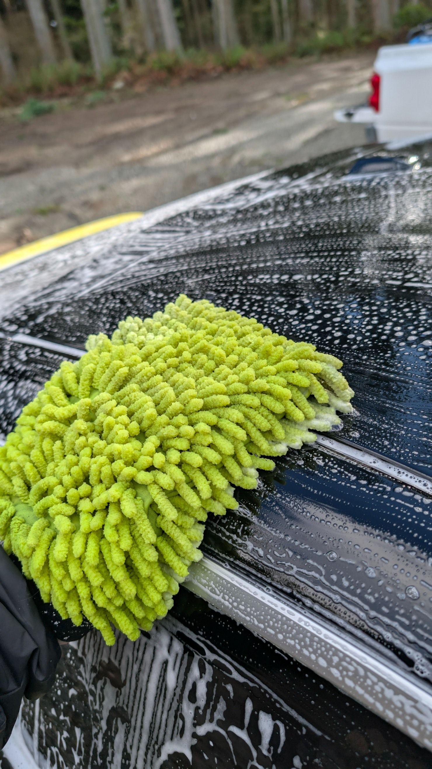 A bright green microfiber wash mitt cleaning soap suds off a black car roof outdoors.
