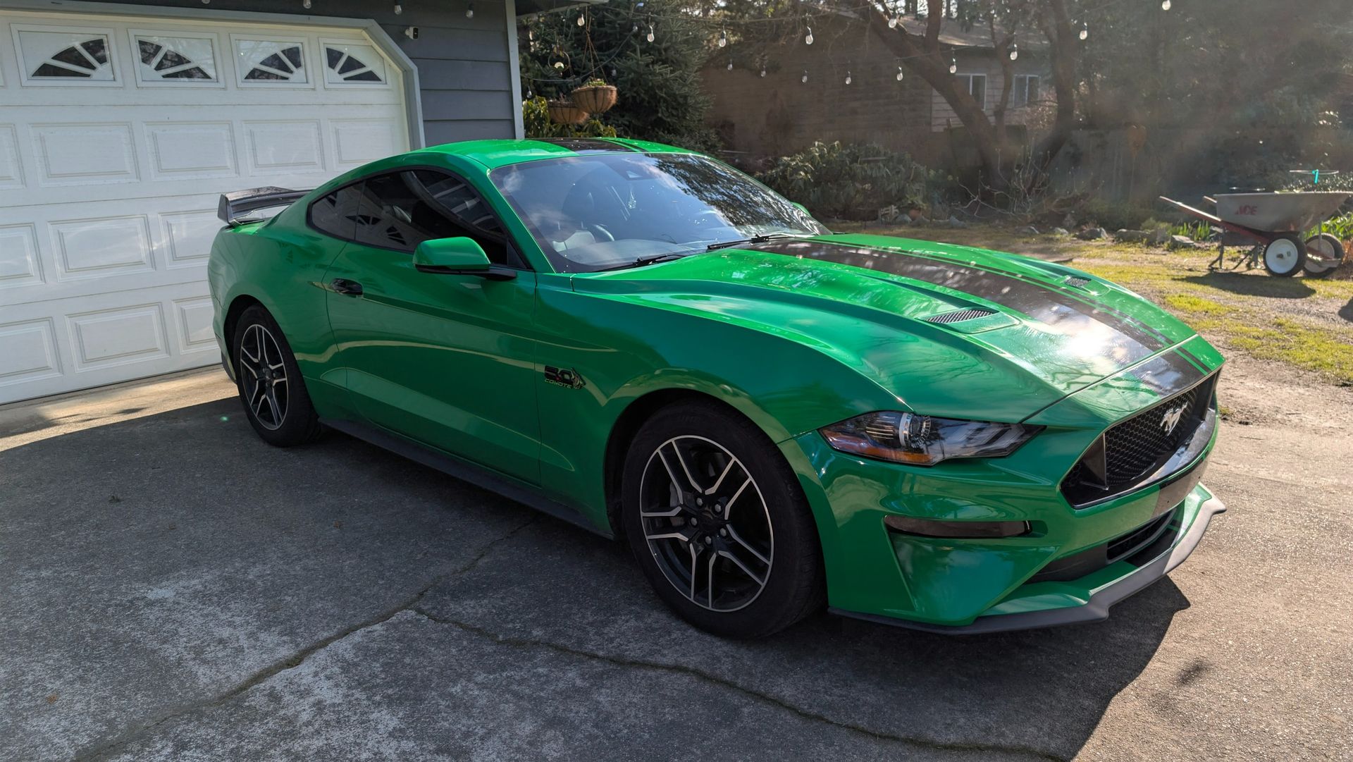 A bright green Ford Mustang with black racing stripes parked on a residential driveway in front of a garage.