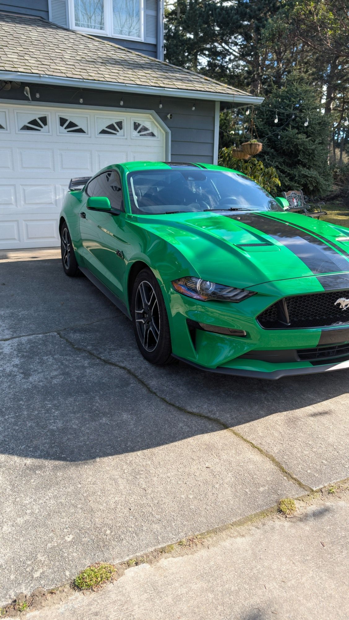 A bright green Ford Mustang with black racing stripes parked in a driveway in front of a house garage.