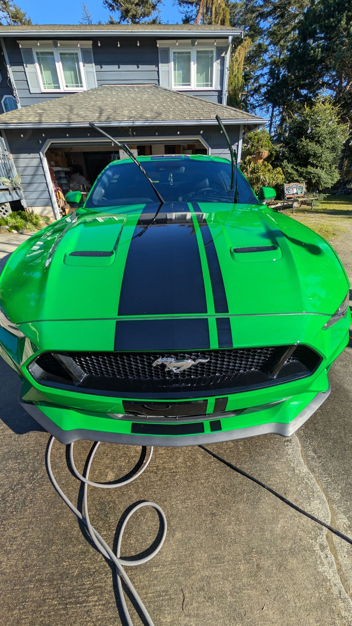 A bright green Ford Mustang with black racing stripes is parked in a driveway in front of a house.