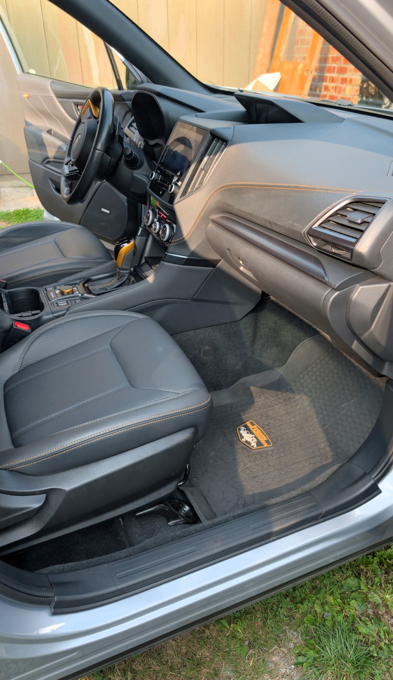 The interior of a grey car showing black leather front seats, a dashboard with a screen, and floor mats.