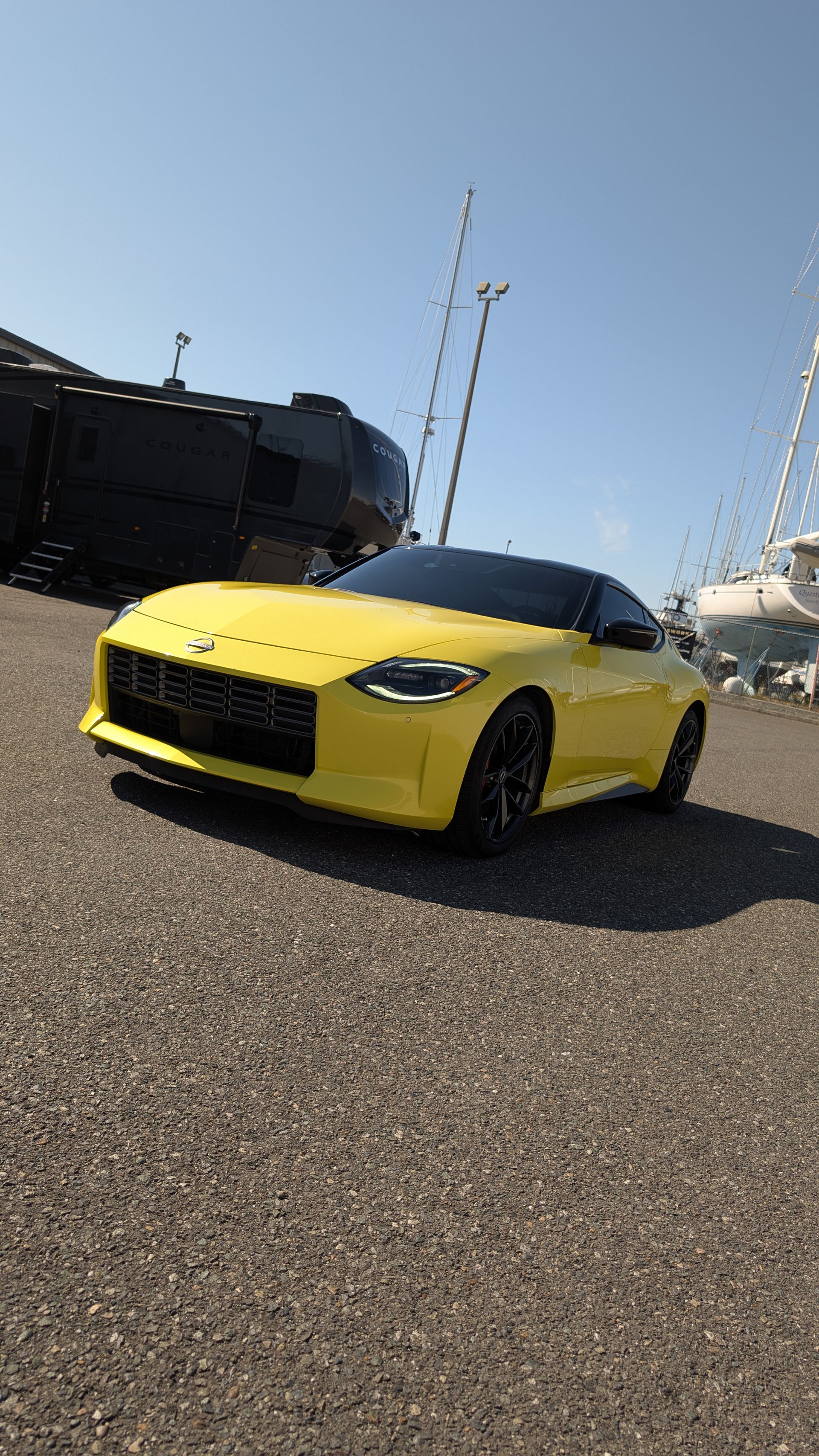 A yellow Nissan Z sports car parked on an asphalt lot with a dark vehicle and boat masts in the background.