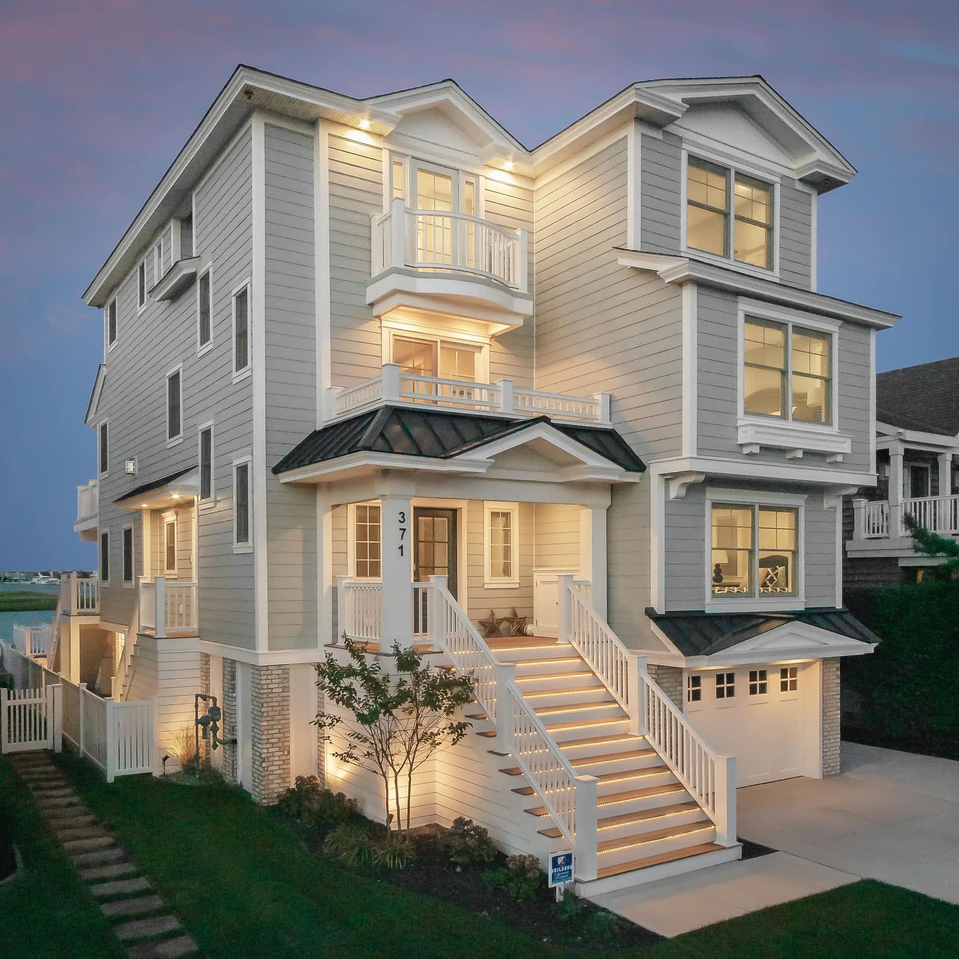 Three-story white house with lit windows, front steps, and a garage at dusk