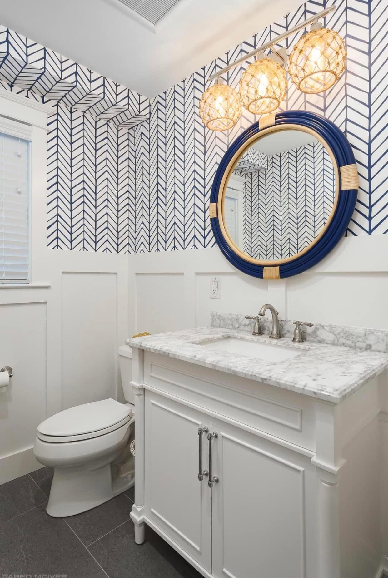 Small white bathroom with navy herringbone wallpaper, round mirror, vanity, and toilet.