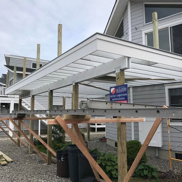 Construction scaffold beside gray house with white roofing beams and support posts on a raised deck.