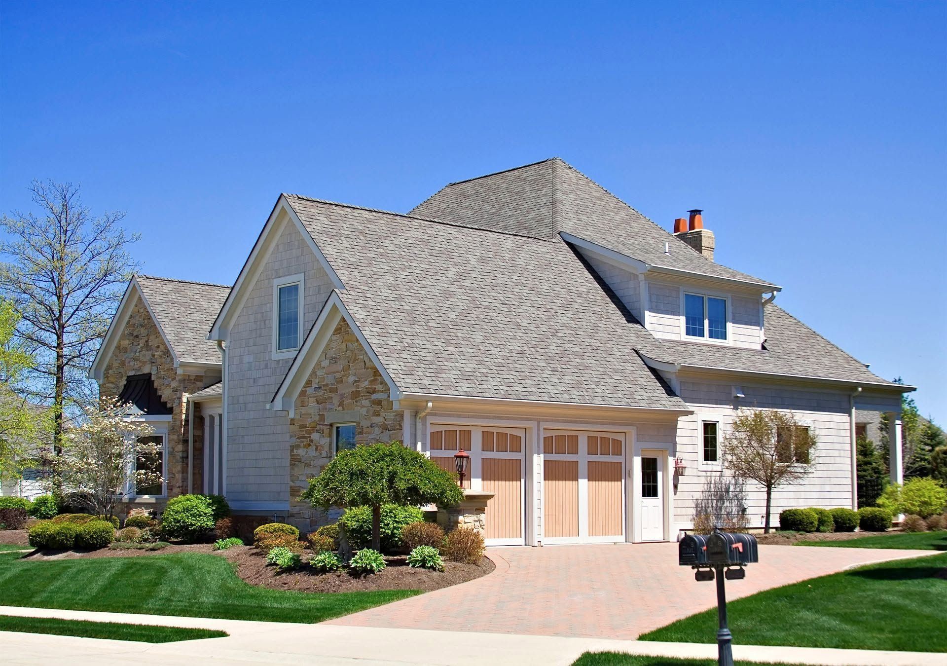 Suburban two-story house with beige siding, gabled roof, and landscaped front yard under a clear blue sky