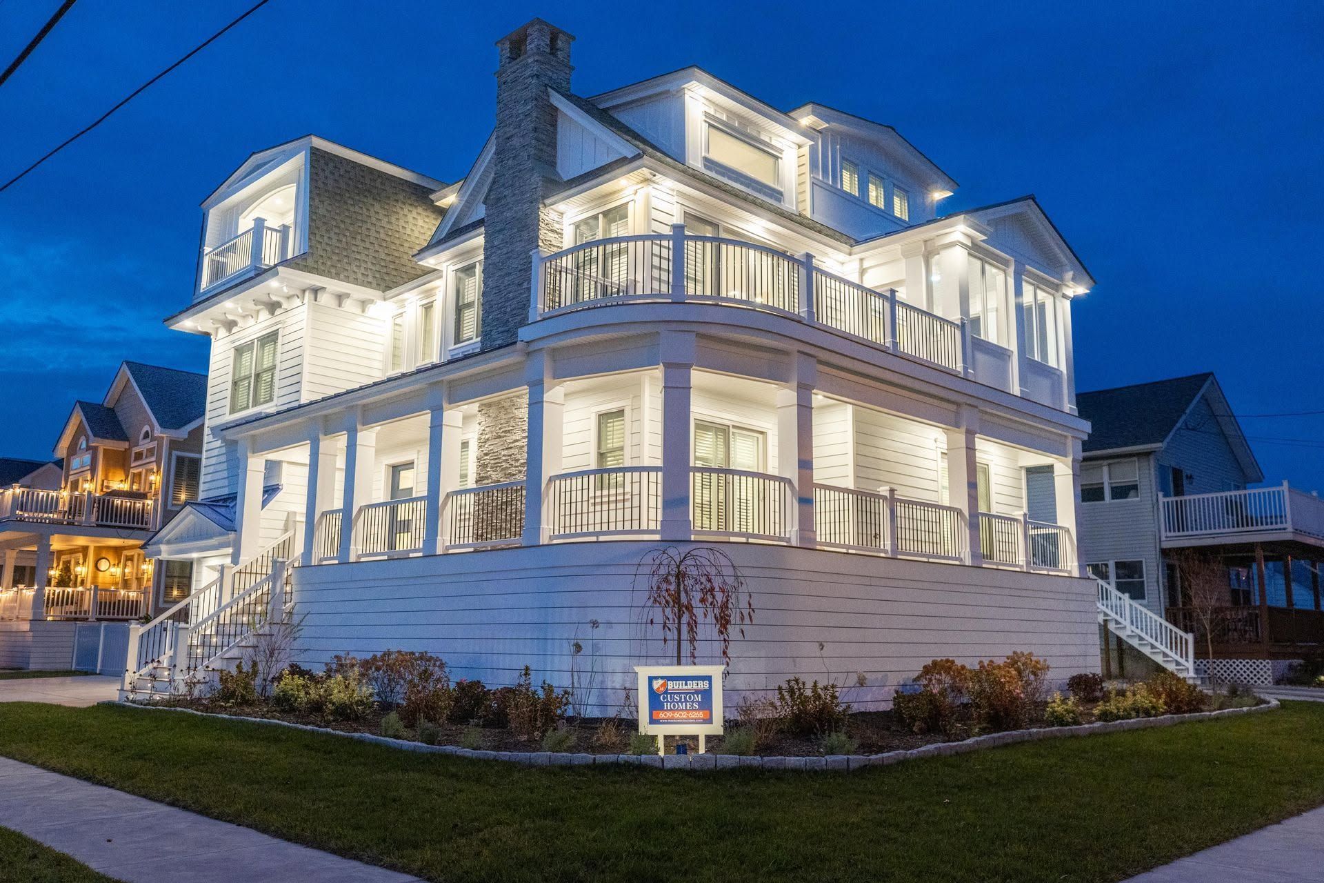 Illuminated white seaside house at dusk with wraparound porches and landscaped yard