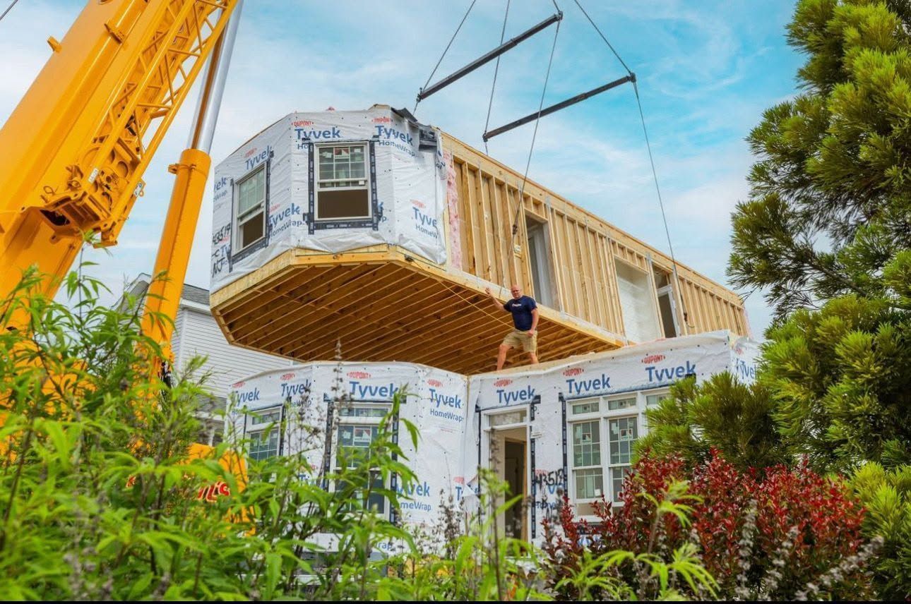 Prefabricated house module being craned into place on a construction site, with trees and blue sky behind it