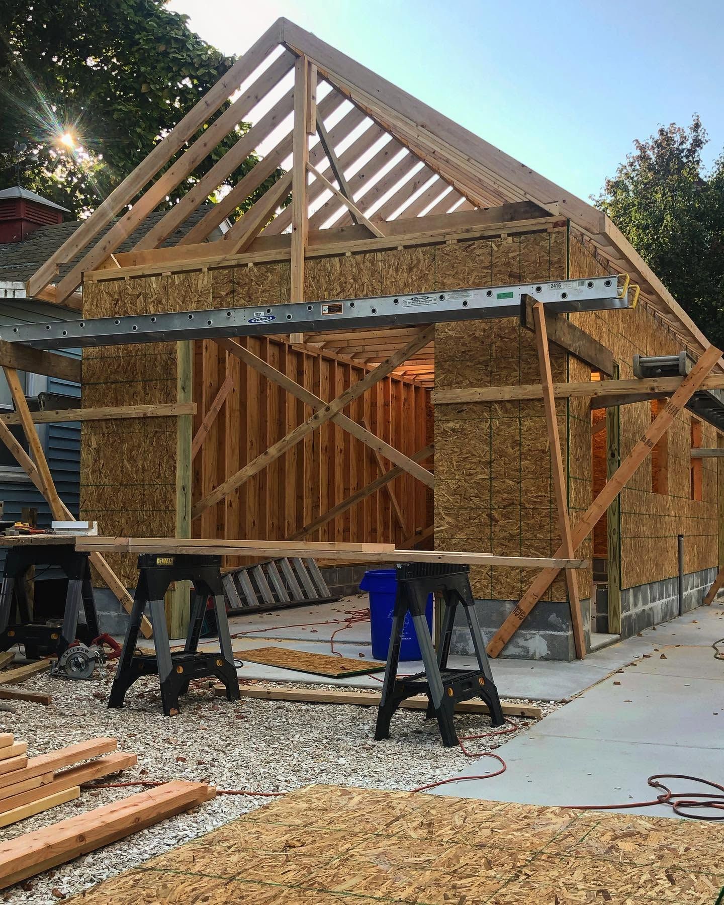 Wood-framed house under construction with roof trusses, sawhorses, and lumber on a gravel driveway