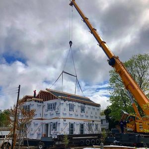 Crane lifting a wrapped modular house onto a trailer at a construction site under cloudy skies