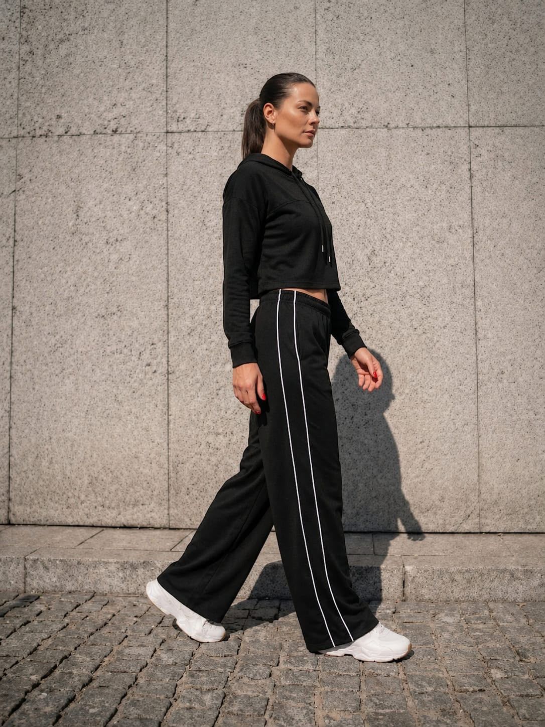 Persona caminando con ropa negra y zapatillas blancas contra una pared gris.
