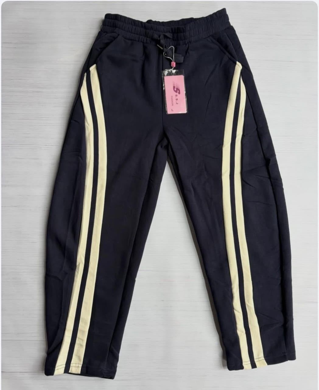 Pantalones deportivos negros con rayas laterales color crema y una etiqueta rosa, extendidos sobre una superficie gris.