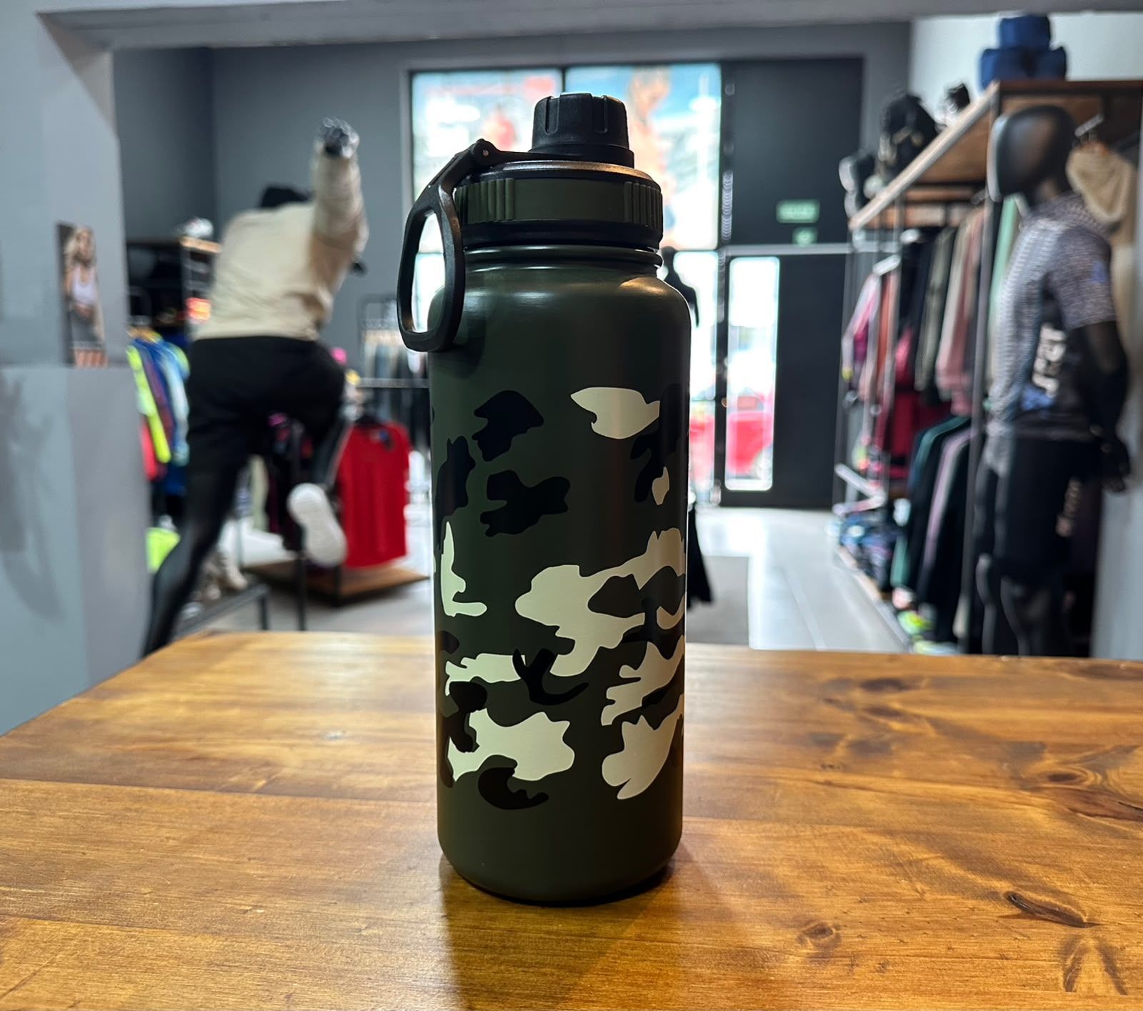 Una botella de agua camuflada sobre una mesa de madera en una tienda de ropa, con percheros y una clienta difuminados al fondo.