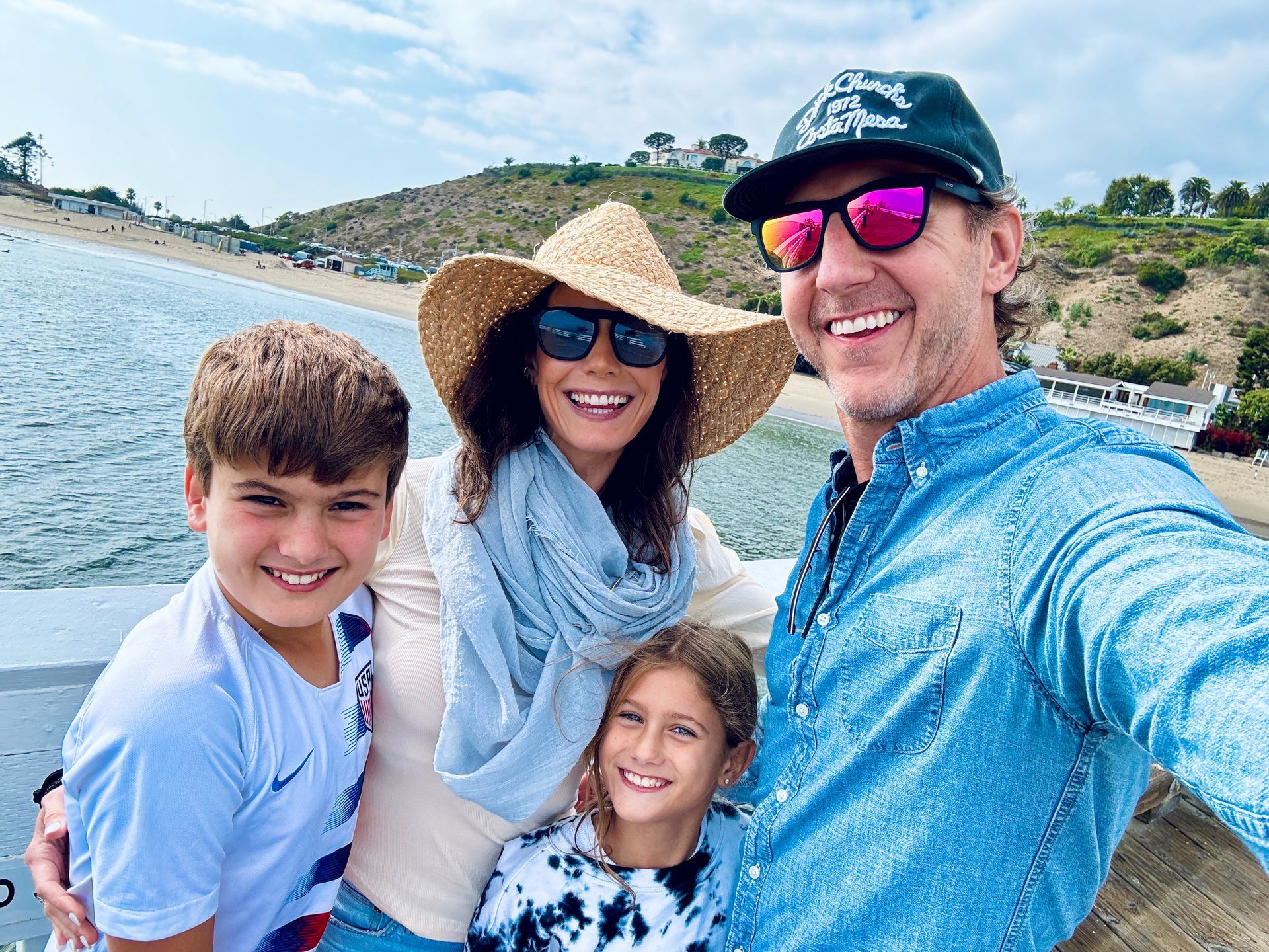 Family selfie on a pier by the beach: Two adults, two children smiling. Sunny day.