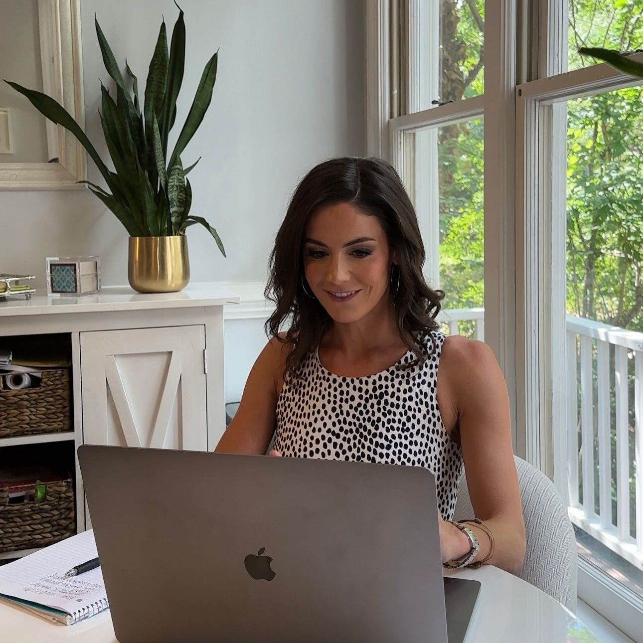 Woman working on laptop at a desk in a bright room. She smiles while looking at the screen.