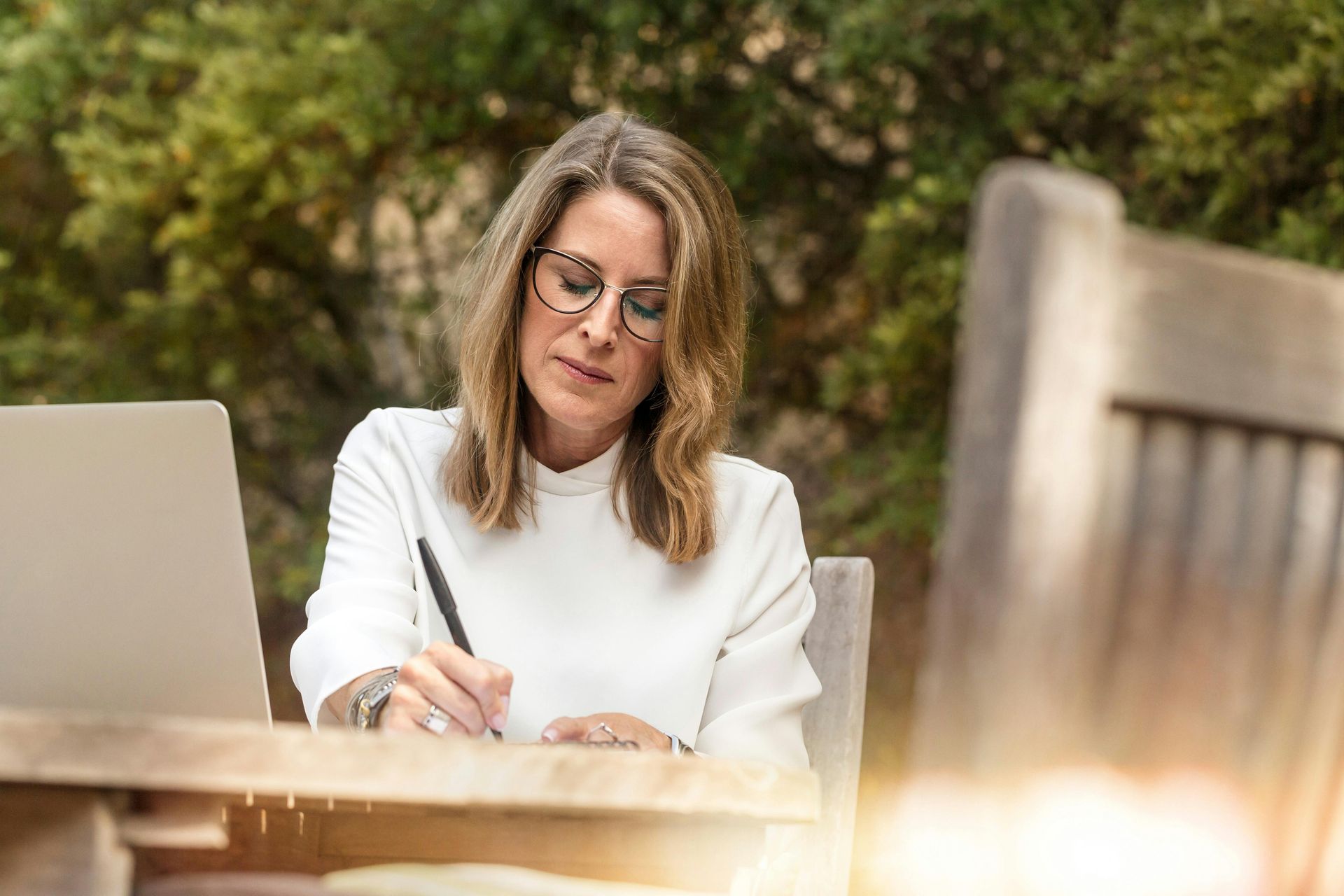 Woman with glasses writing at a table outdoors, laptop nearby, sunny, natural setting.
