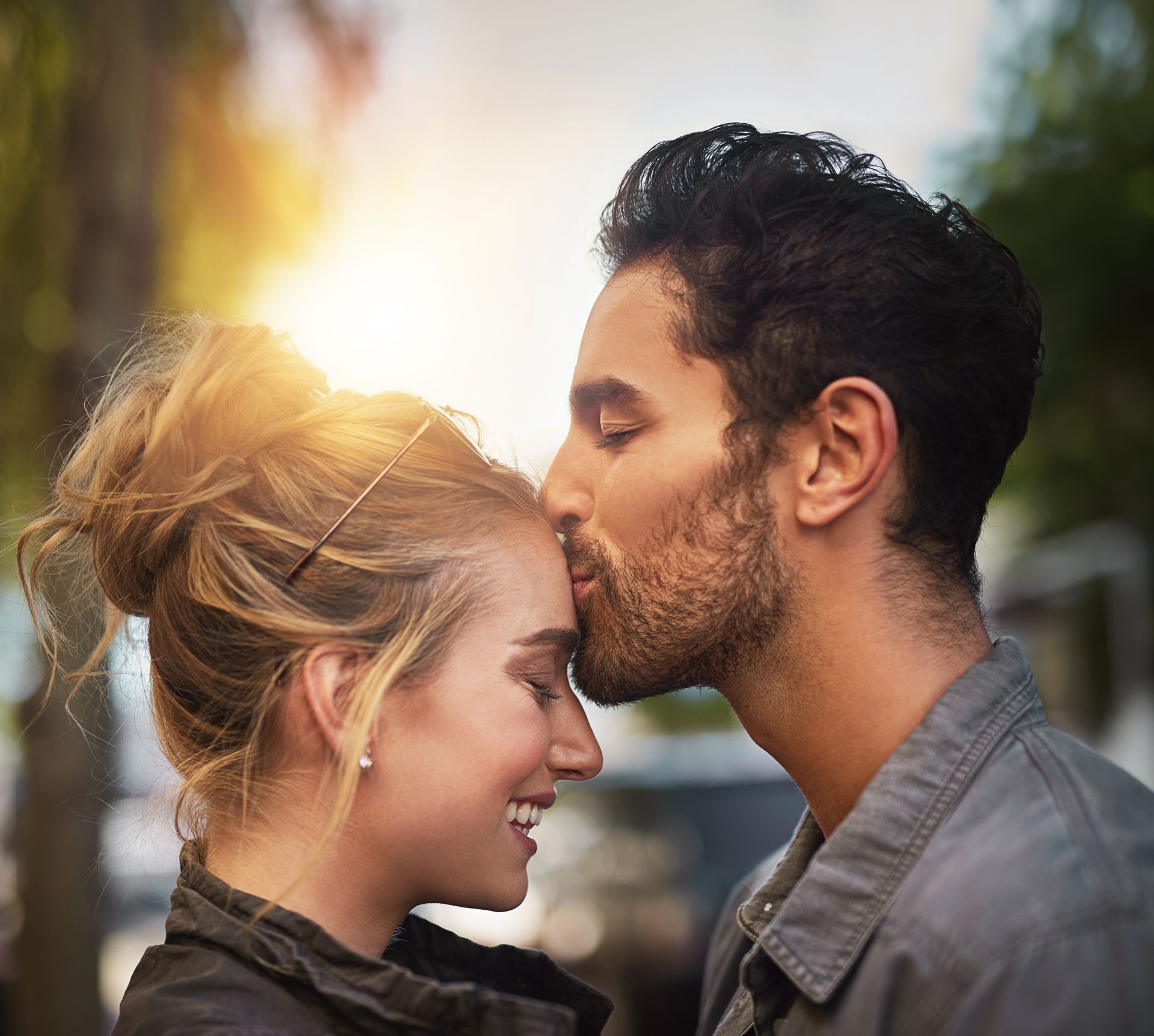 Man kisses woman's forehead; both smile. Outdoors, sunlight, side view.