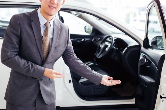 Person in a gray suit presenting an open white car door in a showroom