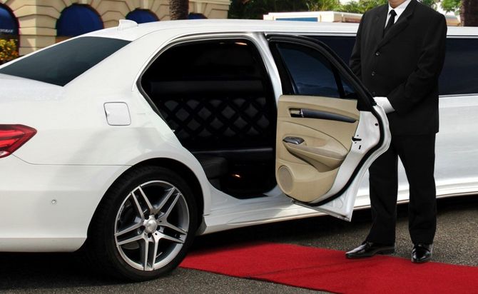 White luxury sedan with open rear door and chauffeur standing beside it on a red carpet.