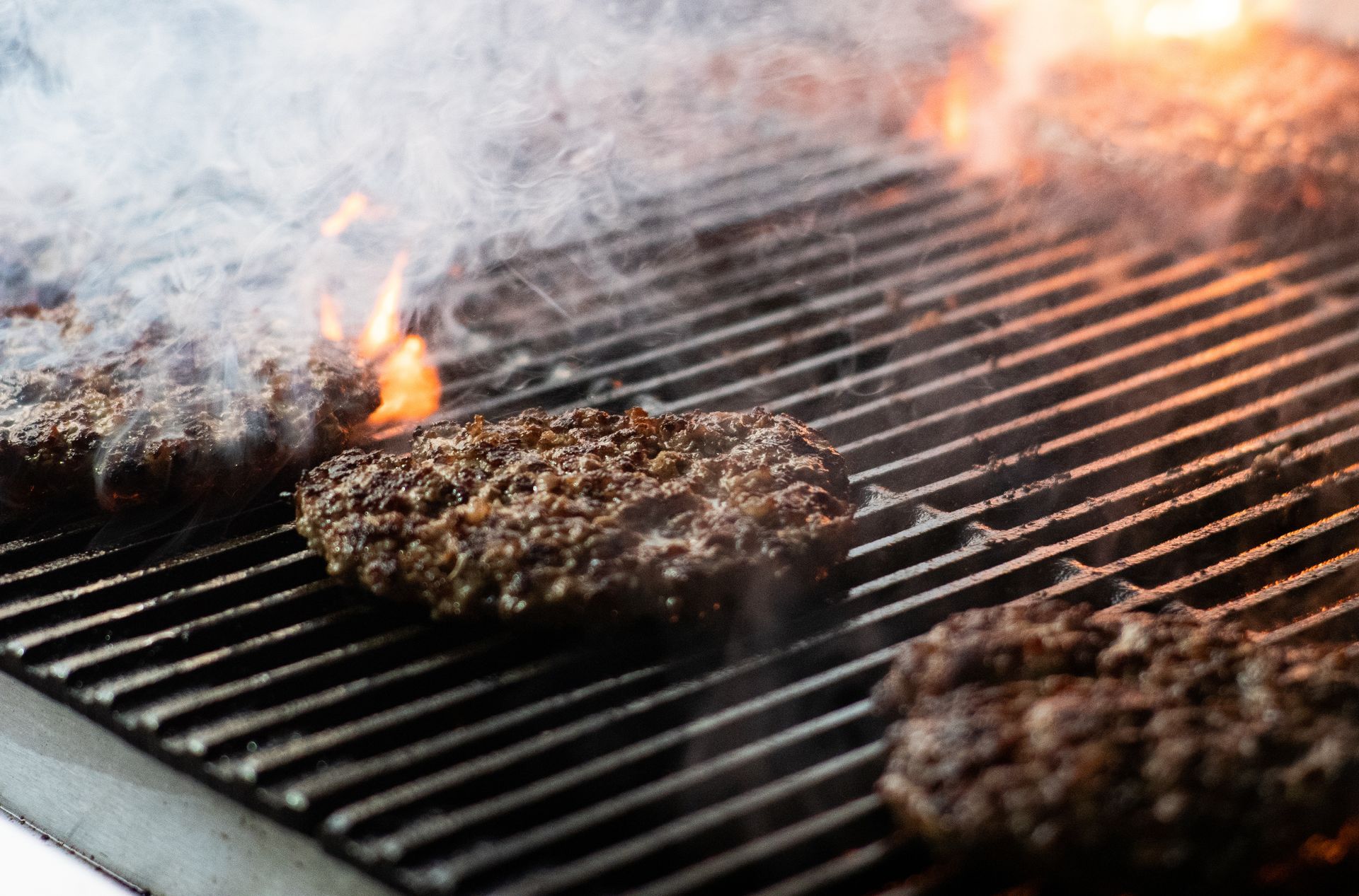 Three hamburger patties are cooking on a grill with flames coming out of it.