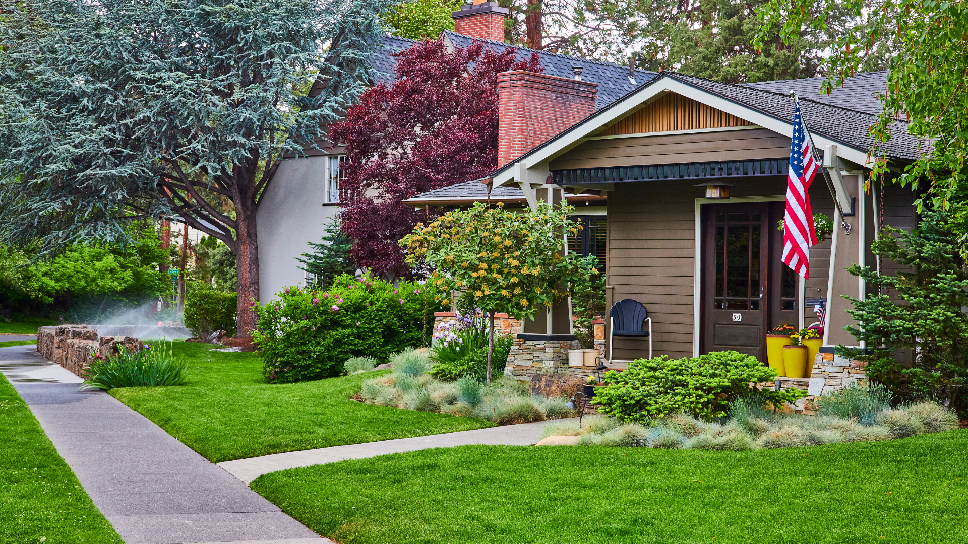 Two-story suburban house with a manicured green lawn, front porch, and trees in the yard.