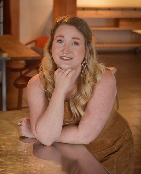 Woman with long blonde hair leaning on a wooden table in a warmly lit room, smiling at the camera