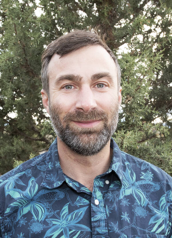 Man standing outdoors in a blue patterned shirt, with trees in the background.