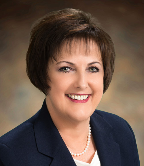 Studio headshot of a smiling woman in a navy blazer and pearl necklace against a blurred brown background