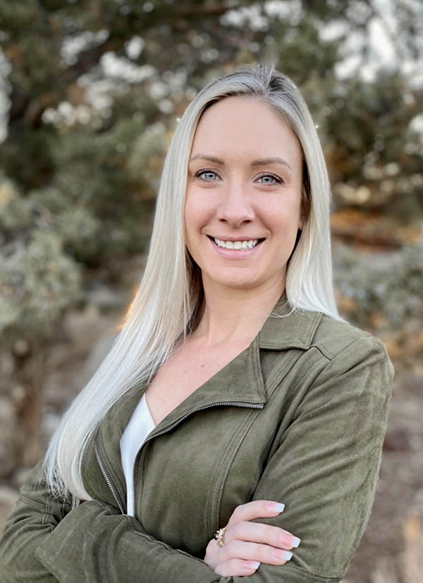 Smiling woman with long blonde hair in an olive jacket, standing outdoors with arms crossed.