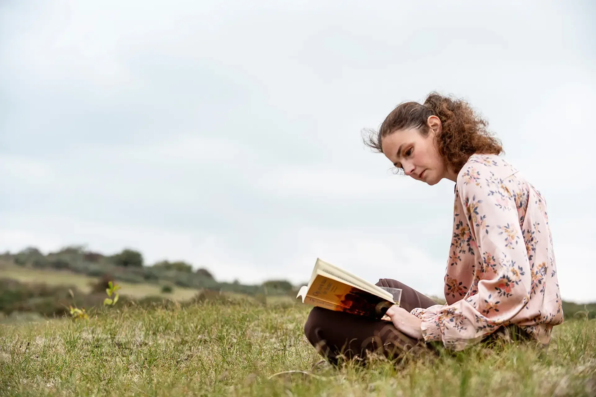 Annemarie zit met haar benen gekruist in een grasveld en leest onder een bewolkte hemel een boek.