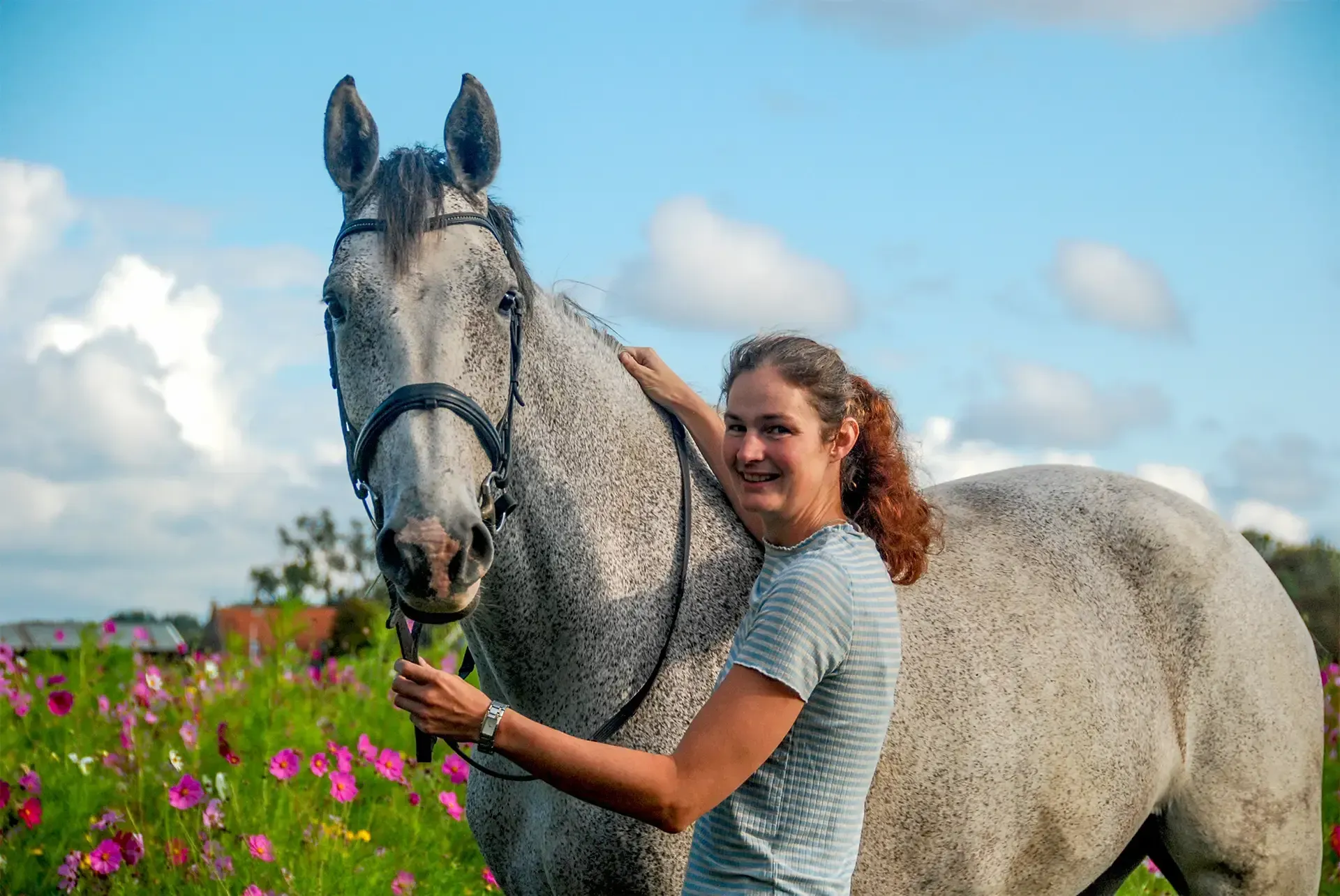 Annemarie omhelst een gevlekt grijs paard in een veld vol roze bloemen onder een blauwe lucht.