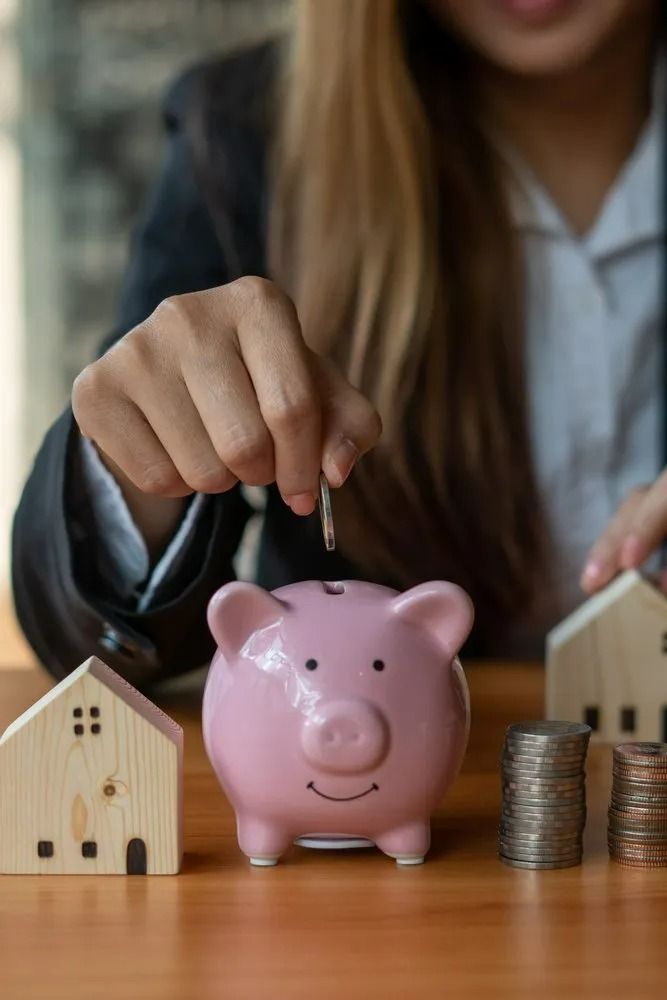 Person Putting Coin in Pink Piggy Bank with Miniature Houses — All Finanz & Insurance Solutions Mackay in Airlie Beach, QLD