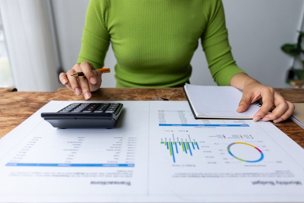 Person in green sweater using calculator, reviewing financial documents, and taking notes at a wooden desk — All Finanz & Insurance Solutions Mackay in Cannovale, QLD