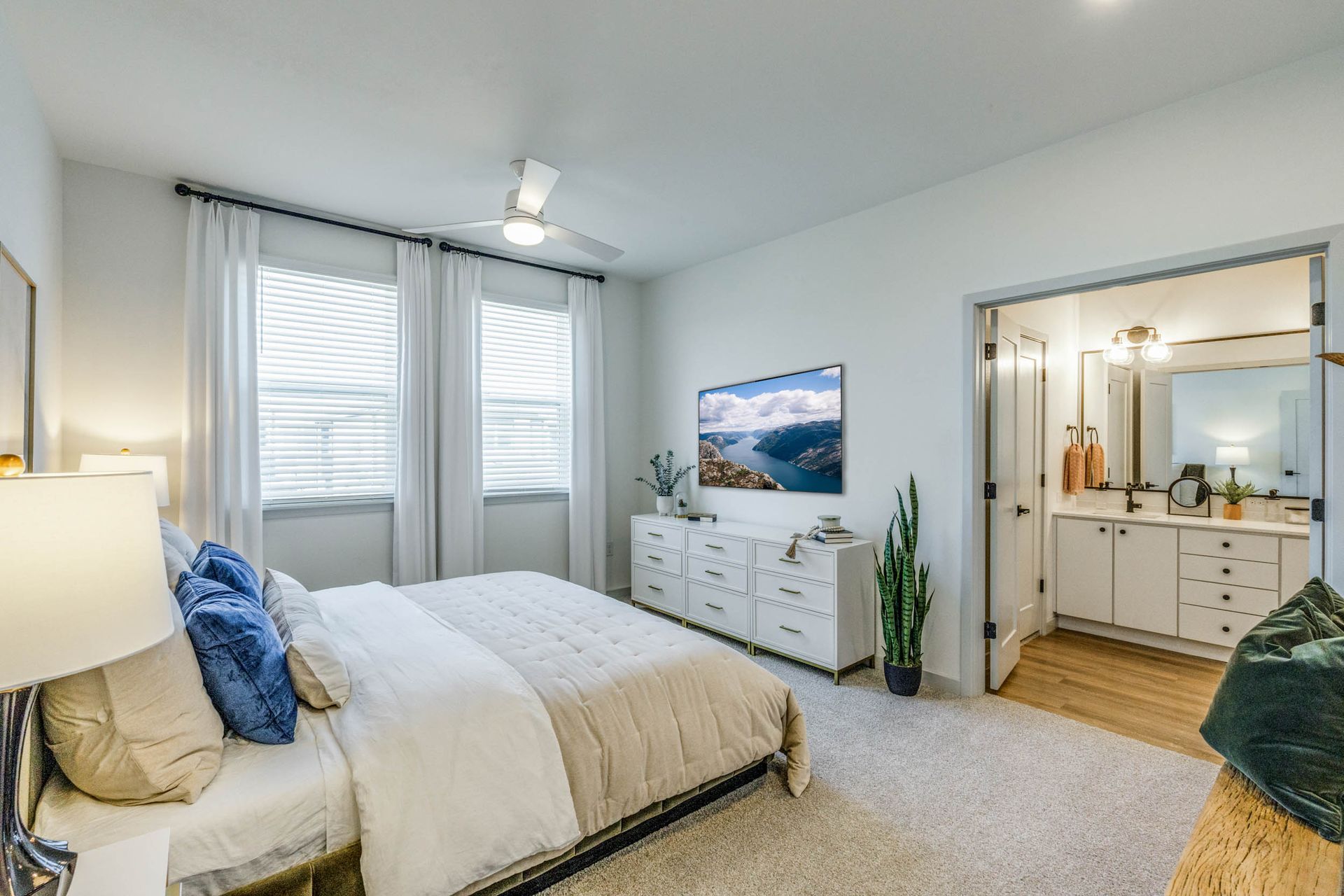 Bedroom with a bed, dresser, TV, and bathroom entry. White walls, beige carpet, natural light.