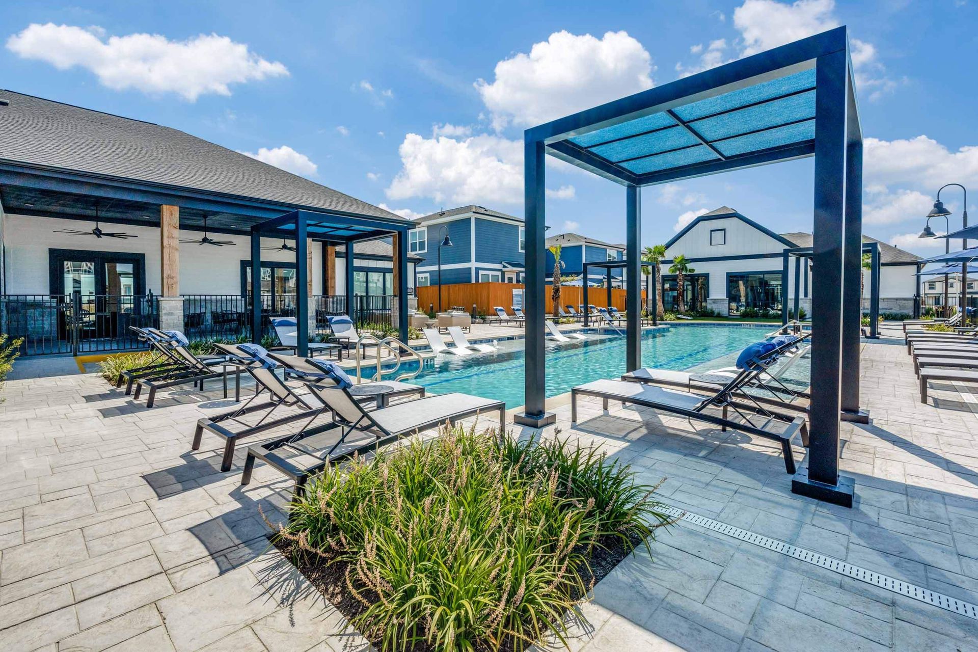 Swimming pool area with lounge chairs and pergola. Buildings in background under blue sky.