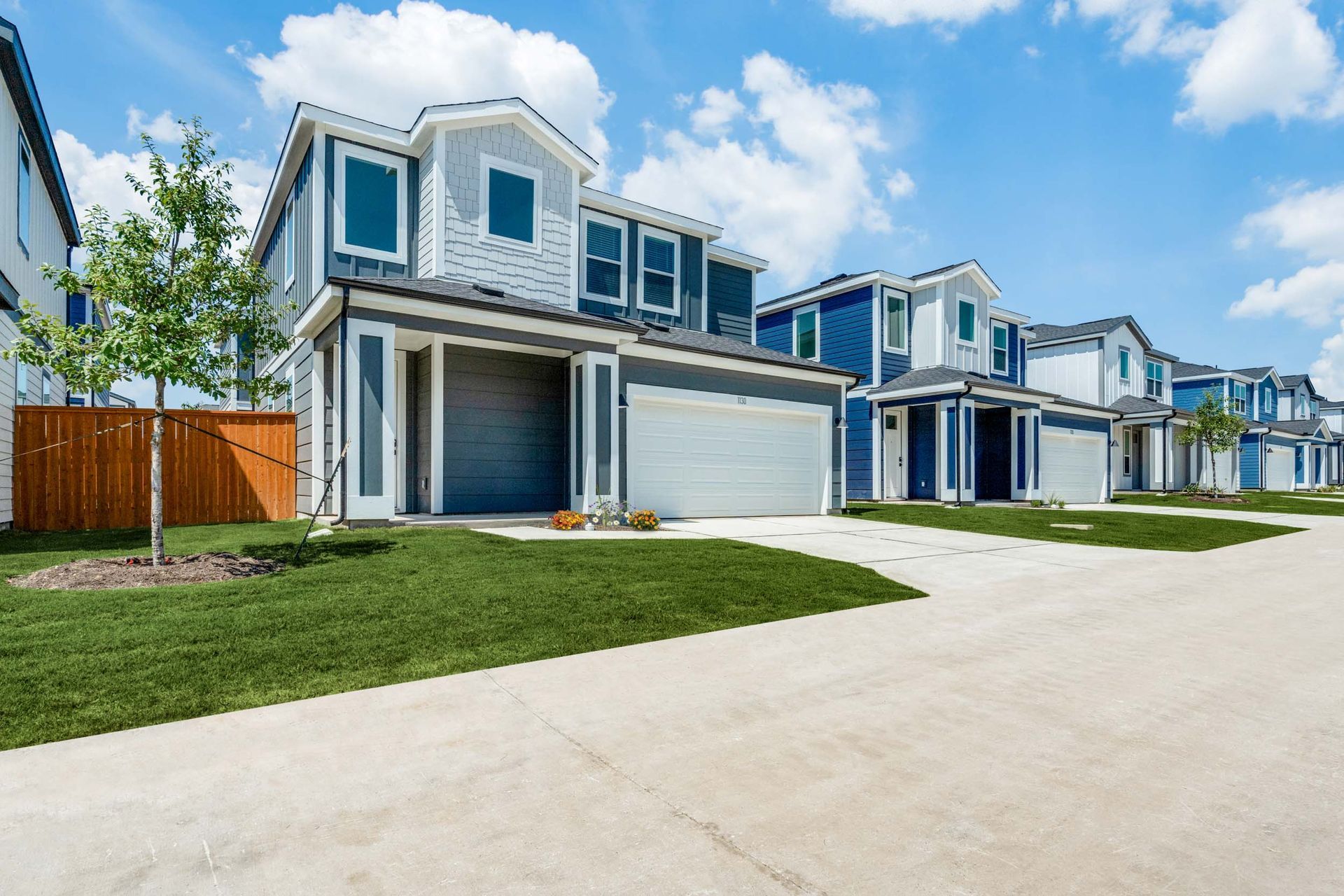 Row of modern townhouses in shades of blue and gray, with garages, green lawns, and a sunny sky.