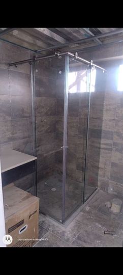 Mampara de ducha de cristal parcialmente instalada en una habitación sin terminar, con las paredes y el techo a la vista.
