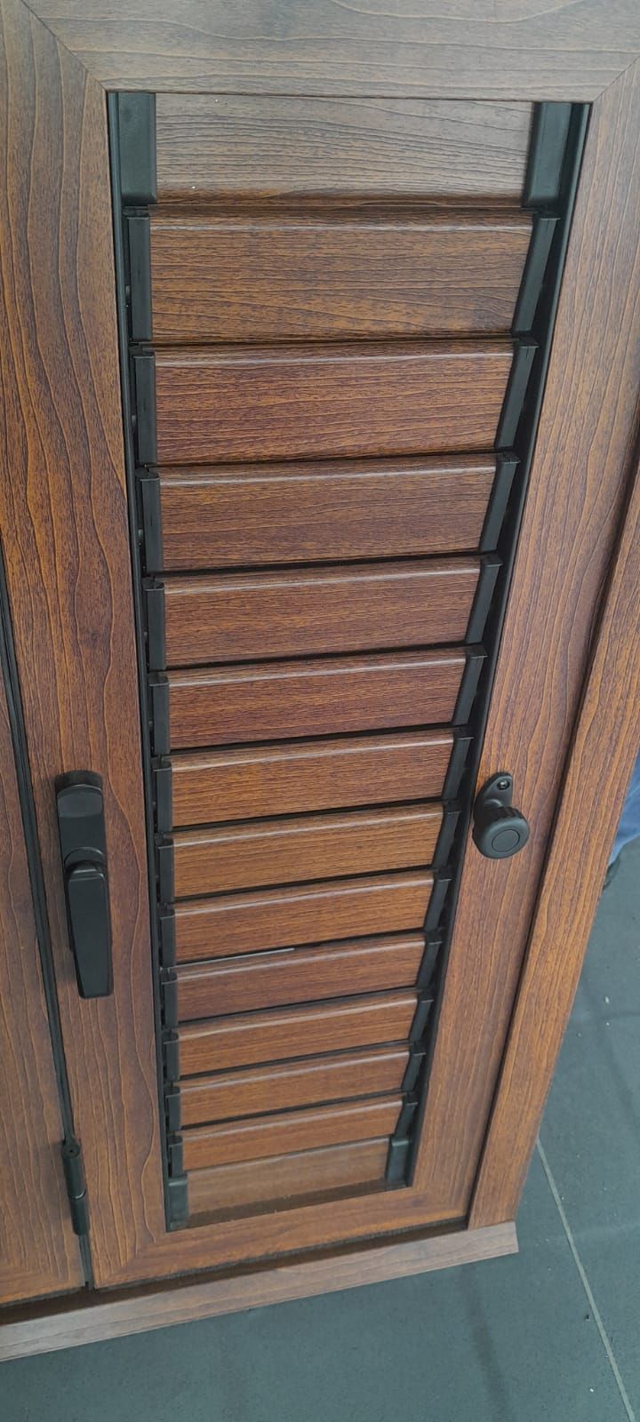 Puerta de armario de lamas de madera, con marco y tirador de metal oscuro, sobre un suelo de baldosas.
