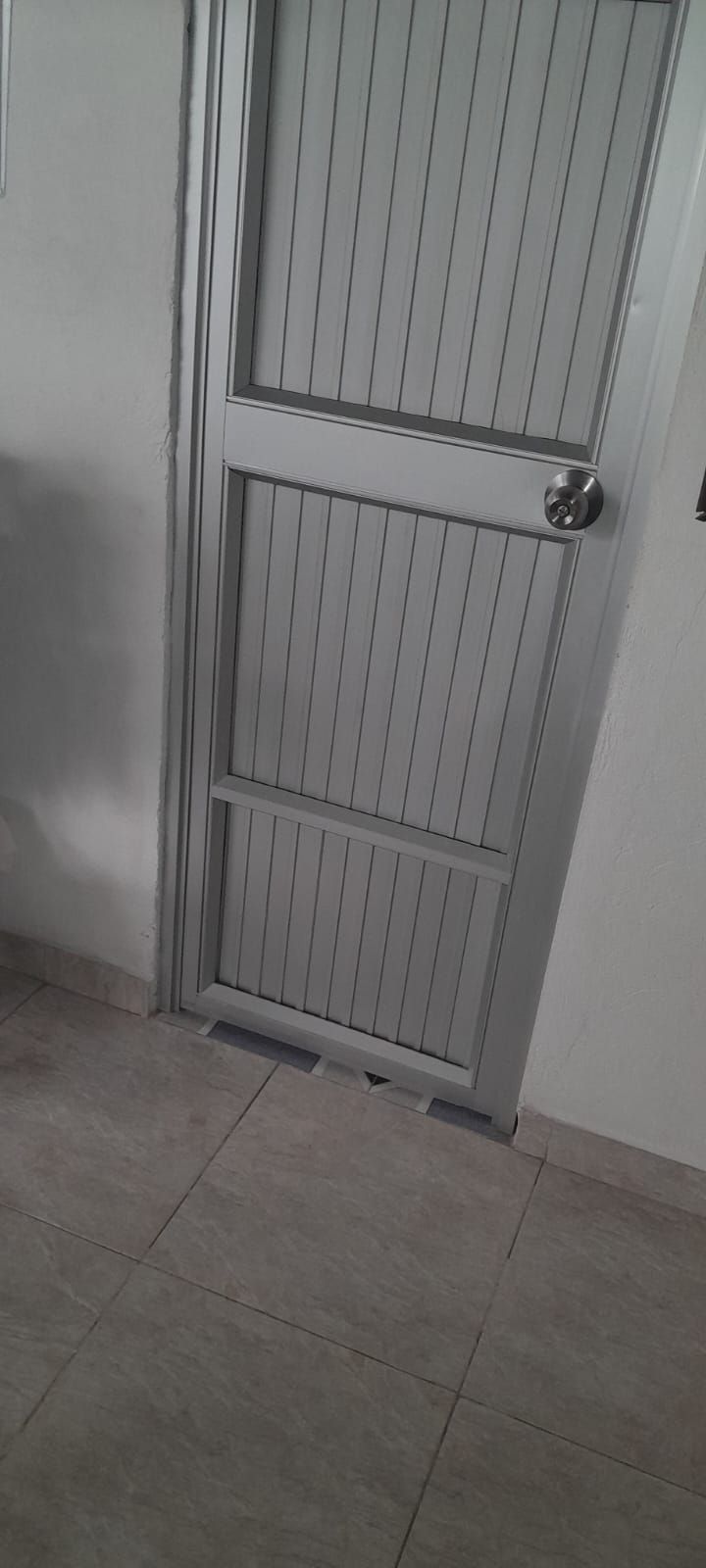 Puerta metálica gris con paneles de listones y un pomo, ligeramente abierta en un pasillo embaldosado.