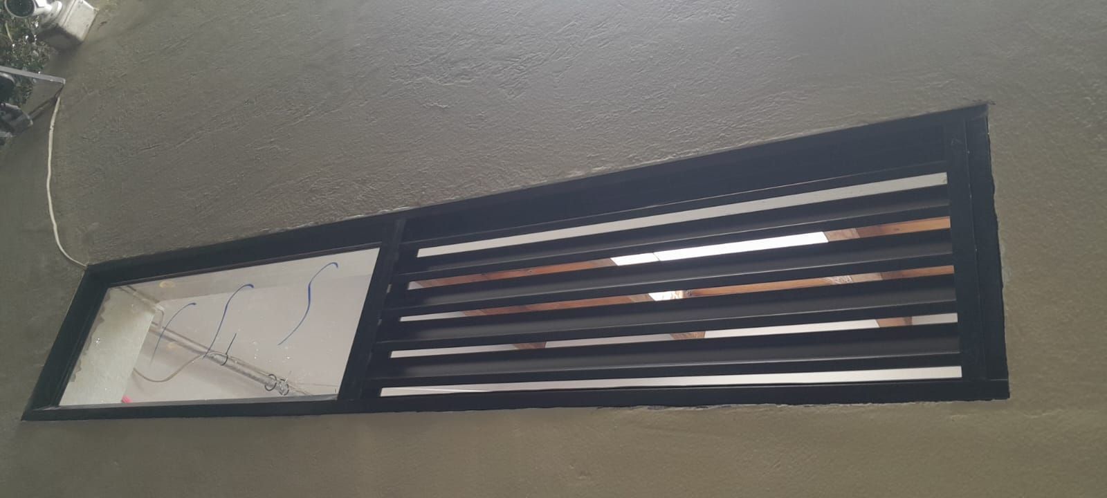 Ventana en un muro exterior con marco oscuro, panel esmerilado y rejilla horizontal de lamas
