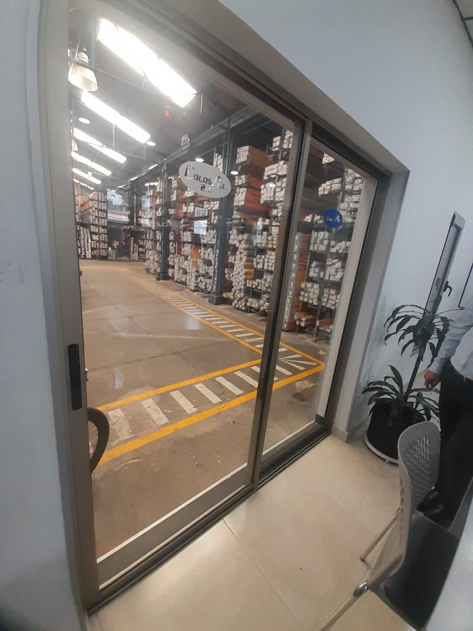 Puerta de cristal abierta que da a un pasillo de almacén con estanterías altas y luces en el techo