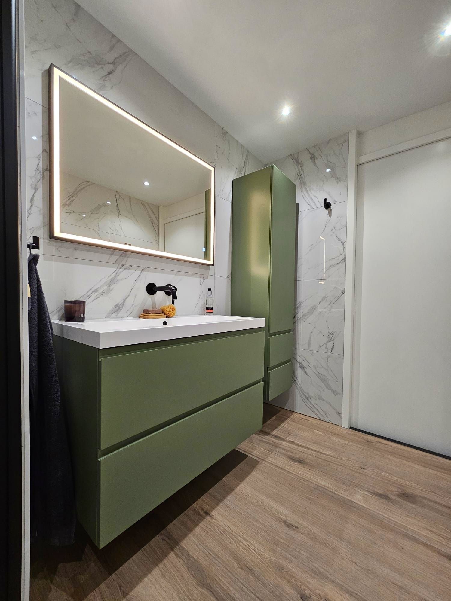 Moderne badkamer met groene wastafelmeubel, verlichte spiegel, marmeren douche en vloer met houtlook.