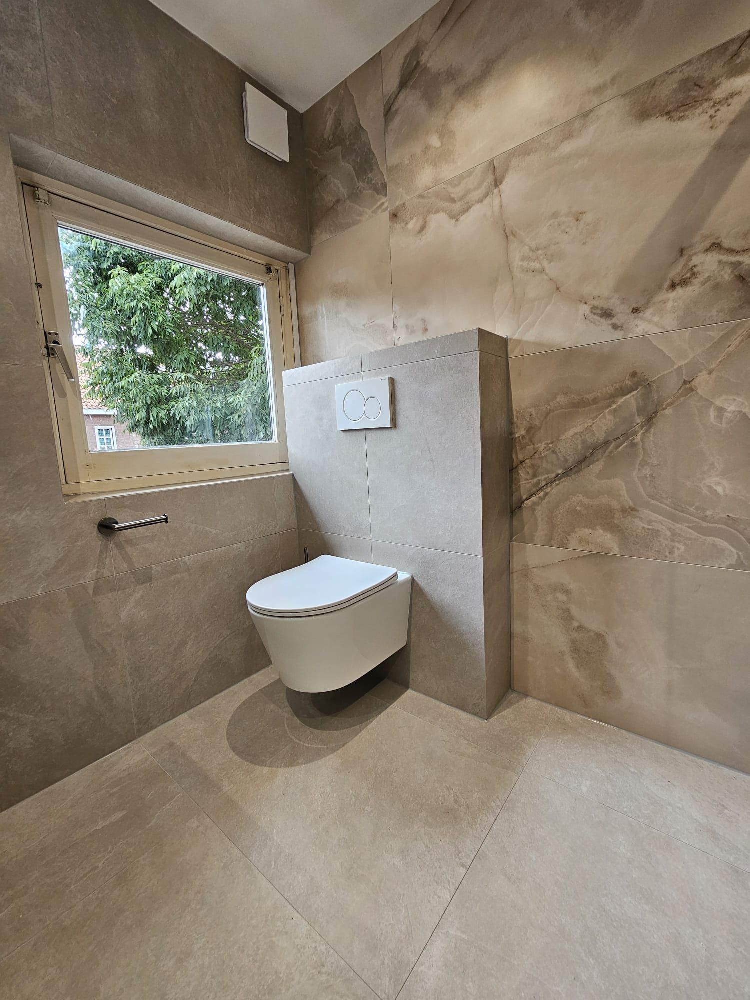 Moderne badkamer met een hangtoilet, beige betegelde muren en een raam met uitzicht op groen