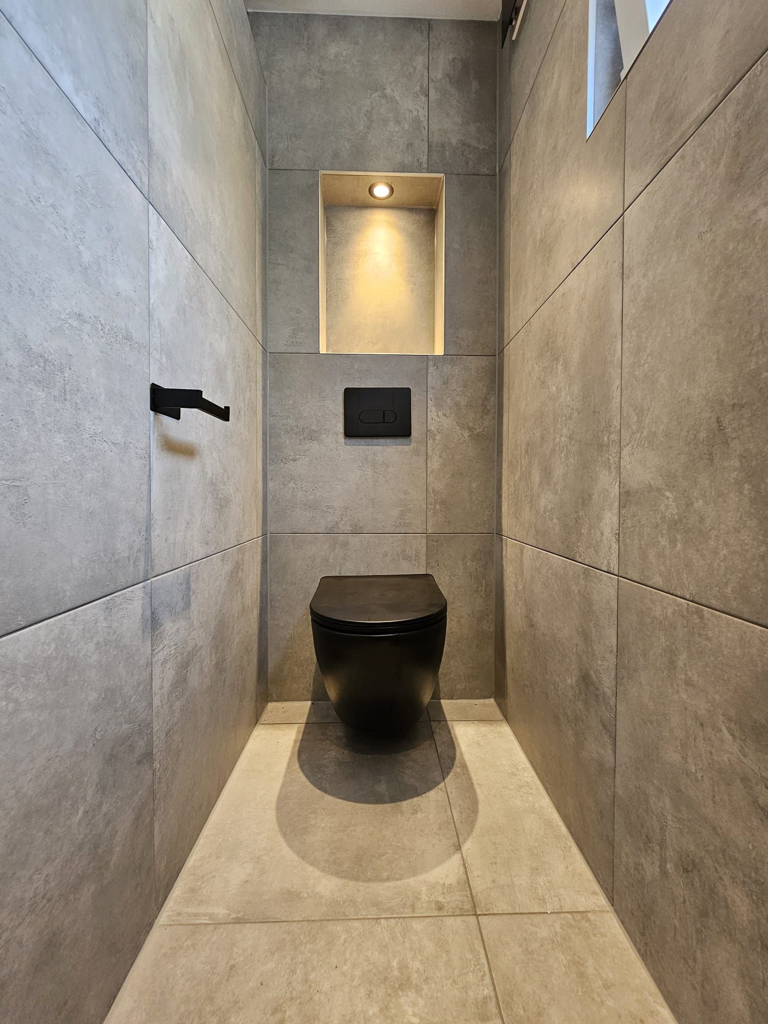 Moderne, smalle toiletruimte met grijze betonnen muren, een hangend toilet en een kleine nis met verlichting boven de wastafel